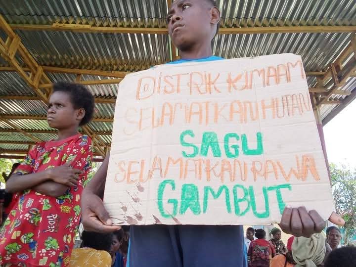 Seorang anak di Kampung Kiworo, Distrik Kimaam sedang memegang sehelai kertas dengan tulisan penolakan PSN saat aksi di Lapangan Maskura-Kimaam – Surya Papua/Frans Kobun